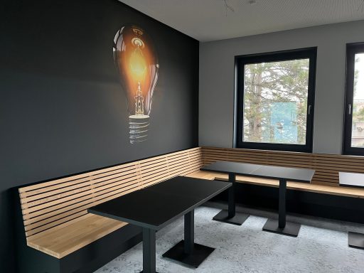 Moderne Cafe-Ecke mit dunkler Wand, Holzbank und Tischen, beleuchtet von einem Wandbild.